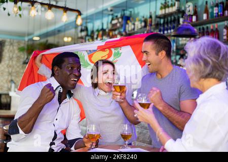 Allegri tifosi multirazziali che sventolano la bandiera del Perù mentre bevono birra e guardano il torneo nello sport bar Foto Stock