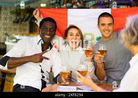 Allegri tifosi multirazziali che sventolano la bandiera del Perù mentre bevono birra e guardano il torneo nello sport bar Foto Stock