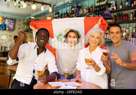 Allegri tifosi multirazziali che sventolano la bandiera del Perù mentre bevono birra e guardano il torneo nello sport bar Foto Stock