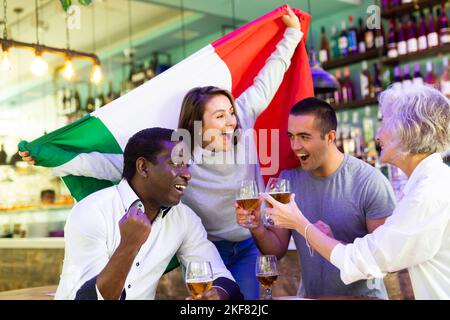 Allegri appassionati di sport internazionali che sventolano la bandiera italiana e bevono birra nello sport bar Foto Stock