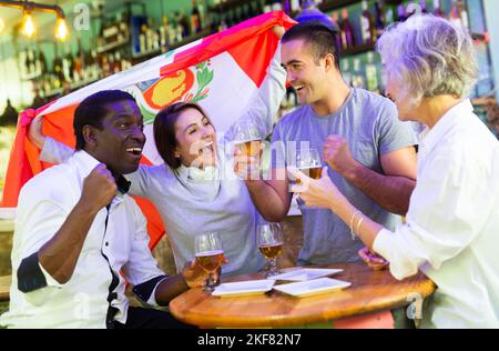 Allegri tifosi multirazziali che sventolano la bandiera del Perù mentre bevono birra e guardano il torneo nello sport bar Foto Stock