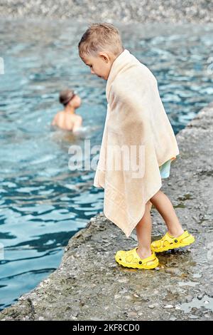 Un ragazzino avvolto in un asciugamano di spugna beige si erge sul molo e guarda l'acqua alla ricerca di una medusa Foto Stock