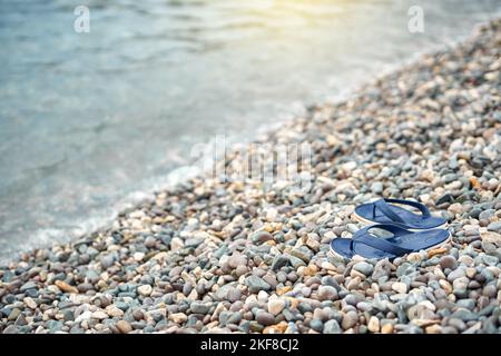 Coppia di infradito blu brillante lasciato sulla spiaggia di ciottoli contro acqua blu sfocata Foto Stock