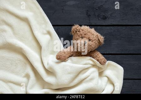 l'orso bruno di teddy si trova sotto una coperta bianca su uno sfondo di legno nero, bambino Foto Stock