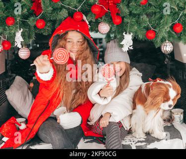 Due ragazze e cane cavalier re charles spaniel con lollipop caramella rossa tra la decorazione di Natale in tronco auto, i bambini hanno divertimento in vacanza invernale Foto Stock