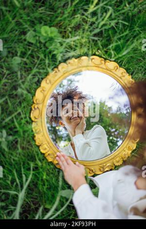 Beautifal donna da corsa mista vestito bianco e riflesso del cielo in specchio rotondo su erba verde primavera. Concetto di natura Foto Stock