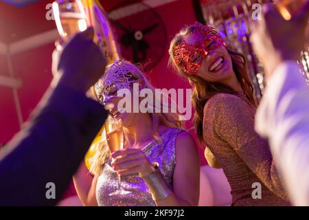 Due donne caucasiche felici che indossano maschere danzanti e bevono champagne ad una festa in un nightclub Foto Stock