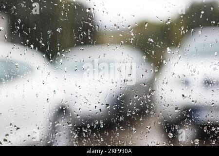 Vetro con gocce di pioggia focalizzate e sfondo sfocato, in un ambiente grigio, nuvoloso, con le auto sullo sfondo. Concetto di pioggia, inverno, tristezza, dus Foto Stock