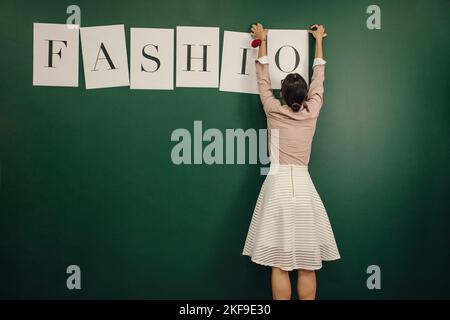 Vista posteriore ripresa di una giovane donna che organizza lettere di parole moda sulla parete. Una designer femminile che si appiccicava la parola sulla parete del suo studio. Foto Stock
