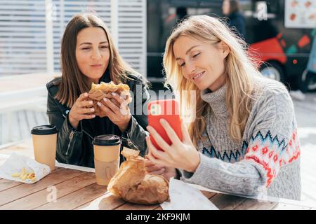 Le donne mangiano il fast food di strada e chiacchierano mentre si siedono al tavolo fuori e usano il telefono per i video. Foto Stock