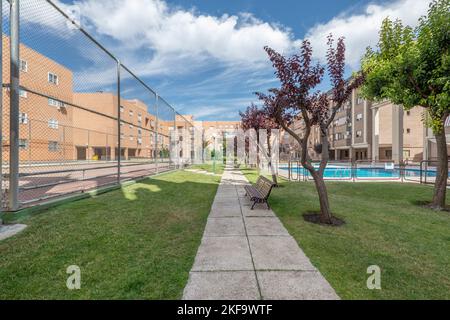 Vista su edifici e aree comuni paesaggistiche con fiori, prati, alberi e piscine estive e un sentiero piastrellato con benche in legno Foto Stock