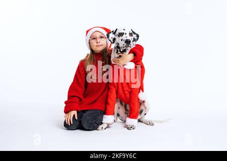 Ragazza carina vestendo maglione a maglia rosso e cappello di babbo natale seduto con il suo amico di animali dalmazia in costume di carnevale di natale. Sfondo, copia Foto Stock