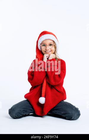 Una giovane bella ragazza che indossa il cappello di Natale siede su un pavimento bianco e guarda la macchina fotografica mentre sorride, tenendo le mani sul suo mento. Isolato Foto Stock