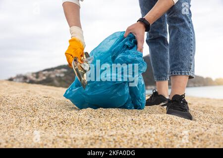 Una donna in guanti con una speciale borsa blu raccoglie rifiuti tra la sabbia lungo la costa. Il problema dell'inquinamento ambientale. Pulizia dei rifiuti Foto Stock