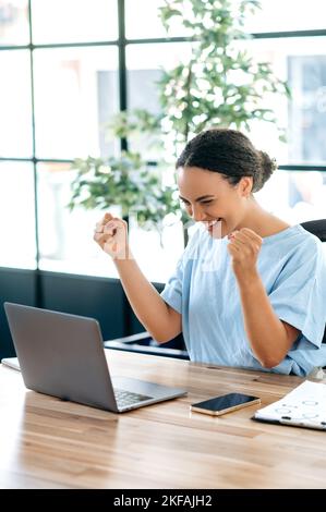 Foto verticale di gioiosa donna ispanica o brasiliana, si siede su una scrivania con un computer portatile in ufficio, gioendo nel successo, grande profitto, celebrare l'affare, gesturing con pugni, guarda il notebook, sorridi Foto Stock