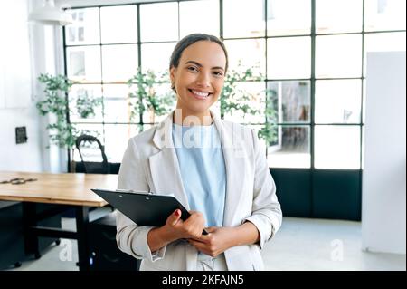 Ritratto di un positivo successo confidente gara mista capelli ricci donna, realtor, top manager, recruiter, tenendo una cartella in mano, si trova in ufficio, guardando la telecamera, sorridendo amichevole Foto Stock