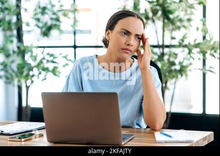 Frustrata stressata triste gara mista giovane donna, impiegato in ufficio, seduto alla scrivania in ufficio, sovraccaricato di lavoro, sentirsi stanco di lavorare in un computer portatile su un progetto, avere un mal di testa, ha bisogno di una pausa Foto Stock