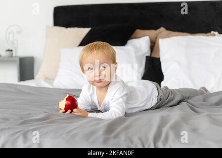 Un bambino carino e sano è sdraiato sul letto. Il bambino tiene e degusta mele sane. Foto Stock