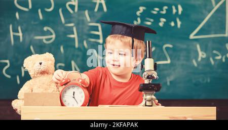 Concetto di pausa scolastica. Il bambino sul viso sorridente guarda la sveglia. Allievo in attesa di una pausa scolastica. Ragazzo in berretto accademico vicino al microscopio, orologio e. Foto Stock