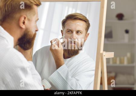 L'uomo adulto focalizzato che si prende cura della pelle con pennello applica crema idratante anti-invecchiamento. Foto Stock