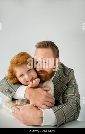 Uomo sorridente in giacca a pipa che abbraccia il figlio rosso e guarda la fotocamera su sfondo grigio Foto Stock