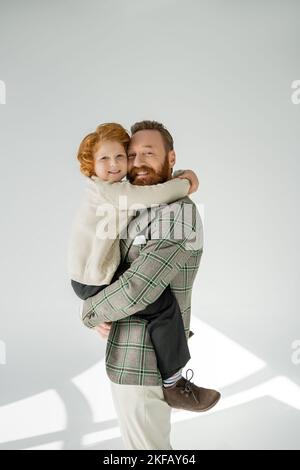 Uomo felice bearded in giacca a scacchi guardando la macchina fotografica mentre abbraccia il figlio su sfondo grigio Foto Stock