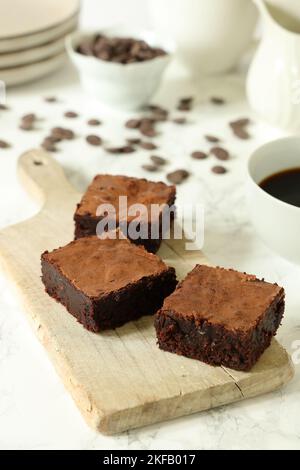 Brownie al cioccolato su un pannello di legno d'annata su sfondo bianco con tazza da caffè, piatti e gocce di cioccolato sparse Foto Stock