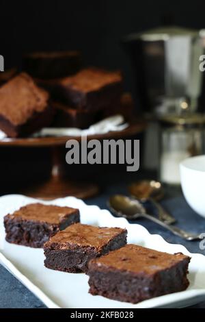 Ricchi e appaganti brownie al cioccolato su un piatto oblungo bianco d'annata su sfondo scuro con base di torta di legno scuro d'annata più caffettiera e tazza Foto Stock