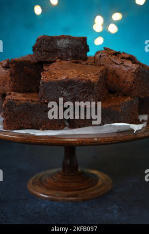 Una pila di ricchi brownie al cioccolato cotti in casa su un piedistallo di torta in legno d'epoca con luci delle fate su un vivace sfondo blu con spazio copia Foto Stock