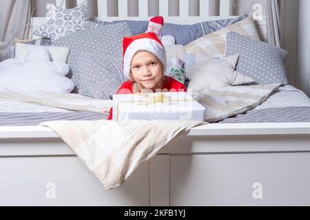 Una ragazza in pigiama rossa e un cappello di Babbo Natale tiene una grande scatola regalo, sdraiata sul letto nella stanza dei bambini. Regali di Natale e Capodanno per i bambini. Famiglia Chris Foto Stock