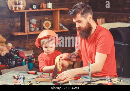 Padre, genitore con barba insegnando al figlio piccolo a segare con affilata sega a mano, carpentiere mestieri. Concetto di doveri maschili. Ragazzo, bambino occupato in protezione Foto Stock