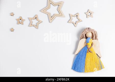 Macrame decorazione natalizia nei colori della bandiera Ucraina. Angelo natalizio e stelle di legno su sfondo bianco. Arredamento fatto a mano. Spazio di copia Foto Stock