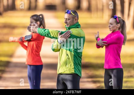 Tre atleti dilettanti, uomo maturo e due giovani donne, si stanno riscaldando prima di fare jogging, facendo stretching della parte superiore del corpo nel parco. Foto Stock