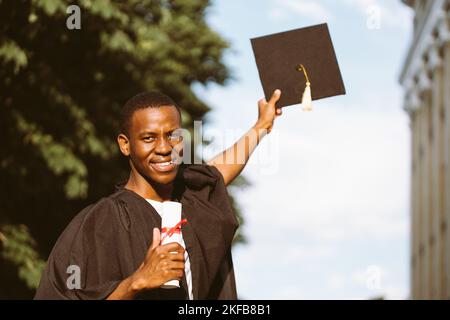 Gioioso afroamericano laureato dall'università stand all'aperto in mantello e tenendo in mano il diploma di istruzione superiore e cappello nero, cielo sfocato Foto Stock