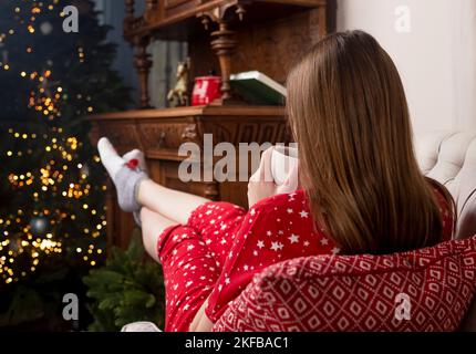 Le gambe femminili con i piedi in calze di Natale carino che si allunga in avanti per l'albero di abete di Natale a casa accogliente e confortevole. Concetto di hygge. Foto Stock