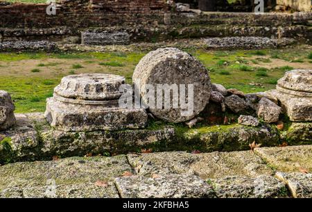Pilastri caduti nelle rovine di Olympia Grecia con antiche pietre di pavimentazione in primo piano e un muro di macerie behind.jpg Foto Stock