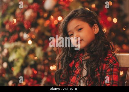 Felice giappone piccola ragazza sognatore guardando via miracolo di attesa da Santa posa vicino a noel albero. Capelli ricci neri asiatici capretto in abito rosso celebrato a plaid Foto Stock