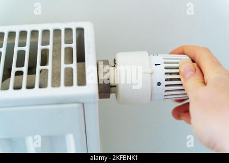 La mano di una donna commuta il regolatore del riscaldamento del radiatore al minimo per risparmiare energia termica a causa dell'aumento dei prezzi per dadi e utenze Foto Stock