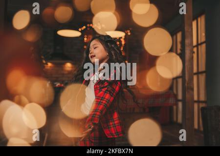 Attiva ragazza bambino giappone con elegante modello di capelli brunette lunga posa a casa in bokeh ghirlands noel. Asian piccolo fashionista in rosso plaid pajama danza Foto Stock