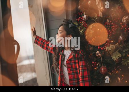 Felice giappone piccola ragazza sognatore guardando attraverso la finestra e miracolo in attesa da Santa posa vicino a noel albero. Bambino asiatico in abito rosso plaid festeggiamento Foto Stock