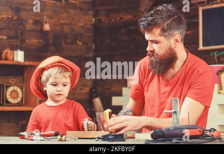 Concetto di paternità. Padre, genitore con barba insegnando al figlio piccolo a segare mentre il figlio gioca con il giocattolo ha visto. Ragazzo, bambino impegnato nell'apprendimento del casco protettivo Foto Stock