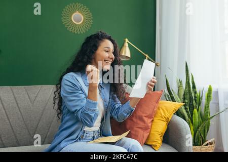 Una giovane ragazza felice studente latino-americano ha ricevuto una lettera con una notifica di esami di successo e l'ammissione all'università. È seduto sul divano, tiene una lettera, legge. Foto Stock