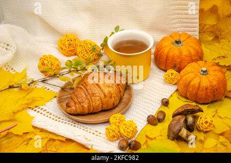 Autunno ancora vita nei toni giallo-arancio: Una maglia bianca e foglie, una tazza gialla e un croissant Foto Stock