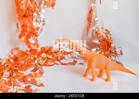Dinosauro giocattolo con collo lungo su sfondo bianco con streamer arancioni. Divertente sfondo orizzontale e spazio copia per gli inviti di compleanno dei bambini. Foto Stock