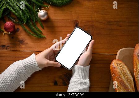 Vista dall'alto, Una donna che tiene in mano uno schermo bianco dello smartphone scherma sopra il suo spazio di lavoro in tema natalizio con sfondo in legno. Foto Stock