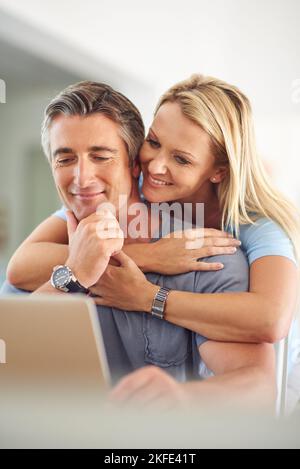 Penso che questo post sul blog sia fantastico. Una donna matura e attraente che abbraccia il suo bel marito maturo mentre usa un computer portatile in casa. Foto Stock