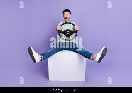 Foto a grandezza naturale di bell'uomo seduto bianco cubo tenere il volante pazzo vestito elegante grigio look isolato su sfondo di colore viola Foto Stock