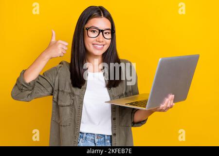 Ritratto di positivo donna ottimista con lunghi capelli indossare camicia cachi mostra come un buon lavoro tenere portatile isolato su sfondo giallo colore Foto Stock