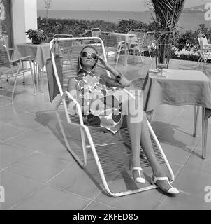 Fashion shoot sull'isola spagnola Ibiza negli anni '1960s. Un modello di moda sulla posizione modella il tipico abito estivo, godendo il sole seduto sulla terrazza dell'hotel. Indossa i grandi occhiali da sole trendy ad arco della decade. 1967 Foto Stock