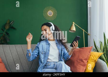 Attraente giovane donna latino-americana ascolta la musica in cuffie bianche, si siede sul divano, danze, tiene il telefono in mano. Foto Stock